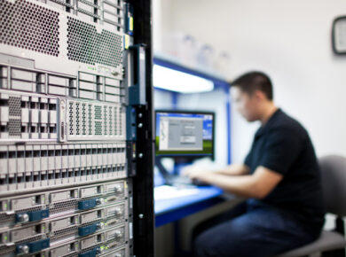 An IT technician is sitting at a workstation next to a tall server rack, watching a monitor in a data centre environment.