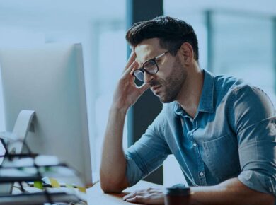 Frustrated man in a denim shirt sits at a desk, hand resting on his forehead while looking at a computer screen.