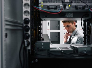An IT professional inspecting a server rack, looking at a laptop in a data centre.