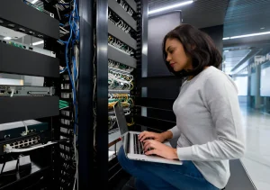 Mujer sentada junto a bastidores de servidores en un centro de datos, escribiendo en una computadora portátil mientras cables de red y equipos se asoman a su alrededor.