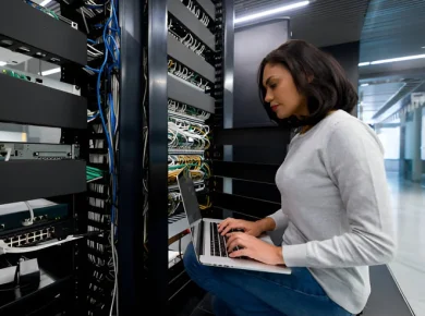 Woman seated by server racks in a data center, typing on a laptop while network cables and equipment loom around her.