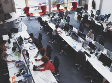 Open-plan office with long desks and multiple computer workstations; employees work at their computers amid large windows and scattered red lounge chairs in the background.