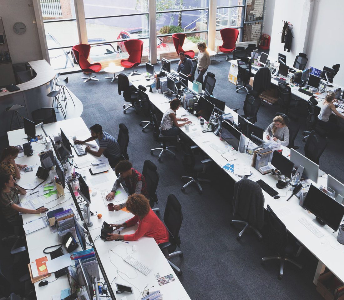 An open-plan office with long desks and several computer workstations; employees work at their computers amidst large windows and a few red lounge chairs scattered in the background.
