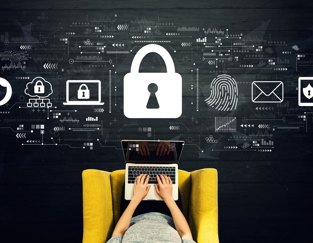 Person typing on a laptop on a yellow chair, with large padlock and digital security icons floating above, illustrating cybersecurity.