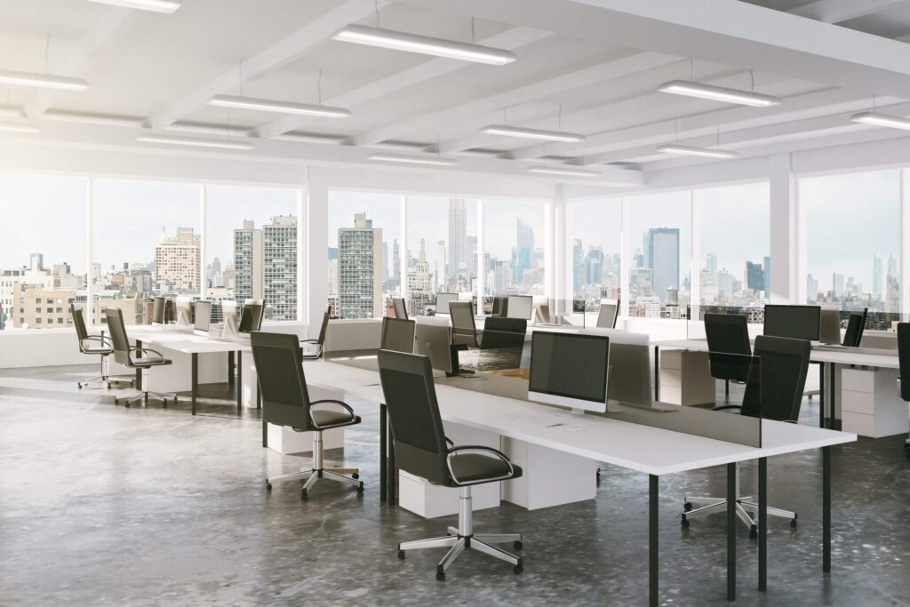 An open-plan office with rows of desks, ergonomic chairs and several computer monitors, looking out over the city skyline through large windows.