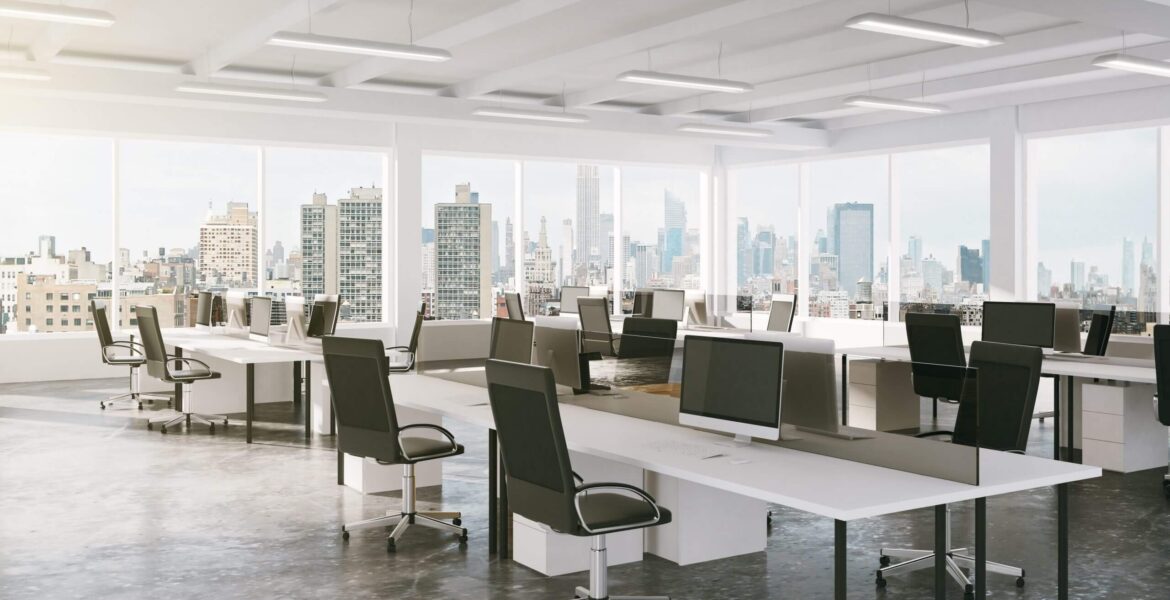Open-plan office with rows of desks, ergonomic chairs, and multiple computer monitors, overlooking a city skyline through large windows.