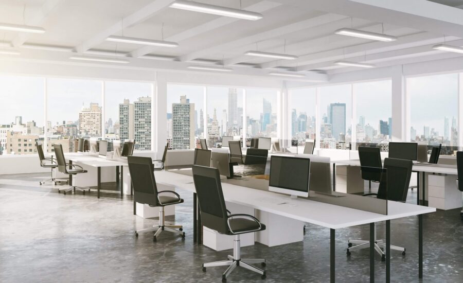 Open-plan office with rows of desks, ergonomic chairs, and multiple computer monitors, overlooking a city skyline through large windows.