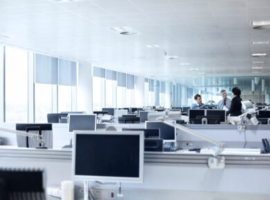 An open-plan office with rows of desks and computer monitors; a small group of three colleagues chatting at the far end.