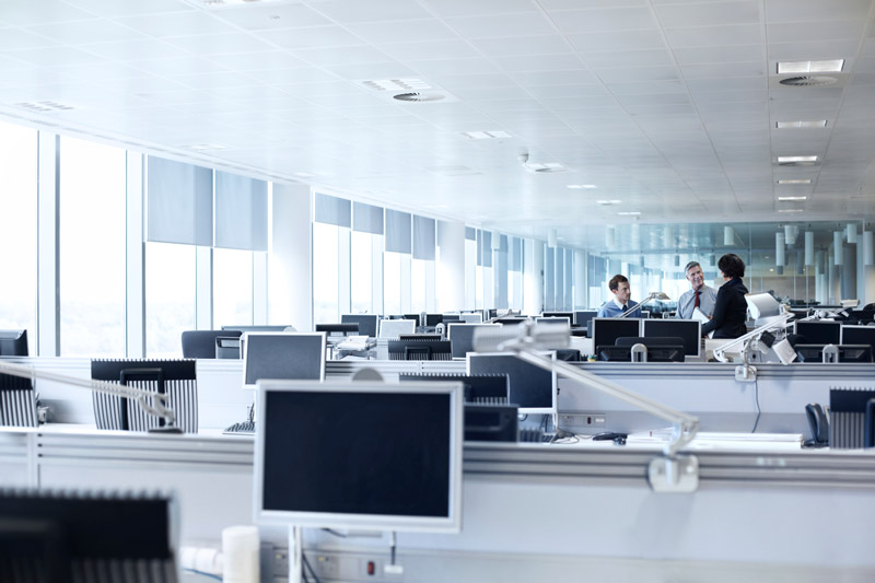 Bureau paysager avec des rangées de bureaux et des écrans d'ordinateur ; un petit groupe de trois collègues discutant au fond.