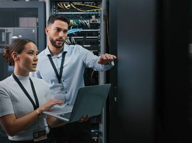 Two IT professionals in a data center review a laptop beside server racks, with one gesturing toward the equipment.