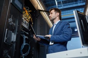 Profesional de TI con traje azul inspeccionando bastidores de servidores con una computadora portátil en un centro de datos