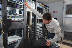 Technician in a data center reaching to rack servers, with a laptop on a rolling cart nearby and network cables in the racks.