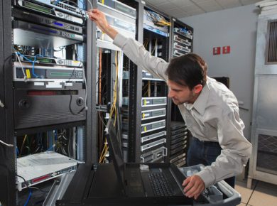 Technician in a data center reaching to rack servers, with a laptop on a rolling cart nearby and network cables in the racks.