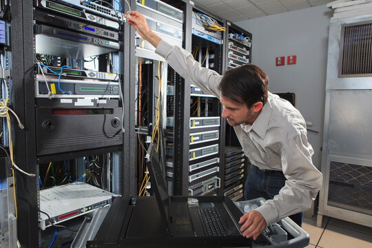 Technician in a data center reaching to rack servers, with a laptop on a rolling cart nearby and network cables in the racks.