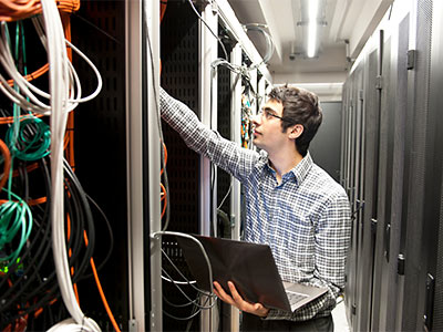 Man in a checkered shirt standing in a server room, holding a laptop and inspecting tangled network cables.