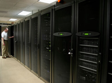 IT technician in a white shirt stands by a row of tall black server racks in a data center corridor, door open to a cabinet.