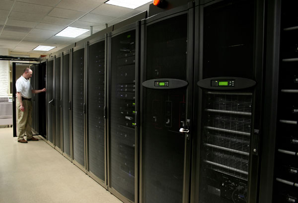 Un technicien informatique en chemise blanche se tient près d'une rangée de hautes baies de serveurs noires dans le couloir d'un centre de données, la porte d'une armoire étant ouverte.