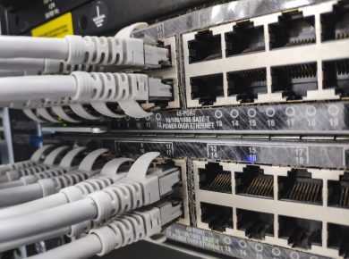 Close-up of a network patch panel with multiple RJ-45 Ethernet ports and gray cables plugged into them in a server rack, showing data cabling.