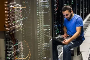 Tech worker in a blue shirt sits beside server racks, using a laptop in a data centre to troubleshoot.