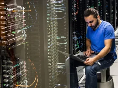 Tech worker in a blue shirt sits beside server racks, using a laptop in a data center to troubleshoot.