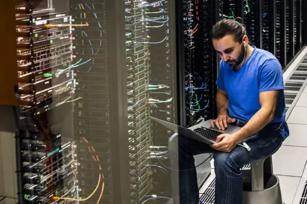 Tech worker in a blue shirt sits beside server racks, using a laptop in a data centre to troubleshoot.